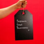 A hand holds a tag with 'Support Small Businesses' message against a vibrant red background.