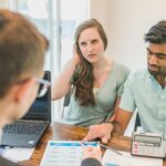 A young couple consults with a realtor about buying a home, indoors in a modern office setting.